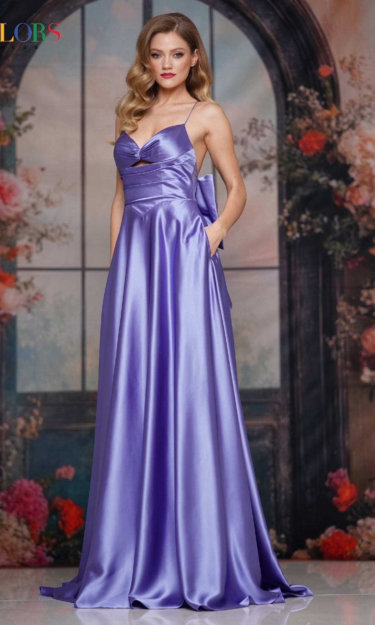 A woman poses before a floral backdrop in the Colors Dress 3851 Back-Bow Strappy Prom Gown, a shiny lavender floor-length evening dress with a sweetheart neckline and thin strappy-back straps.